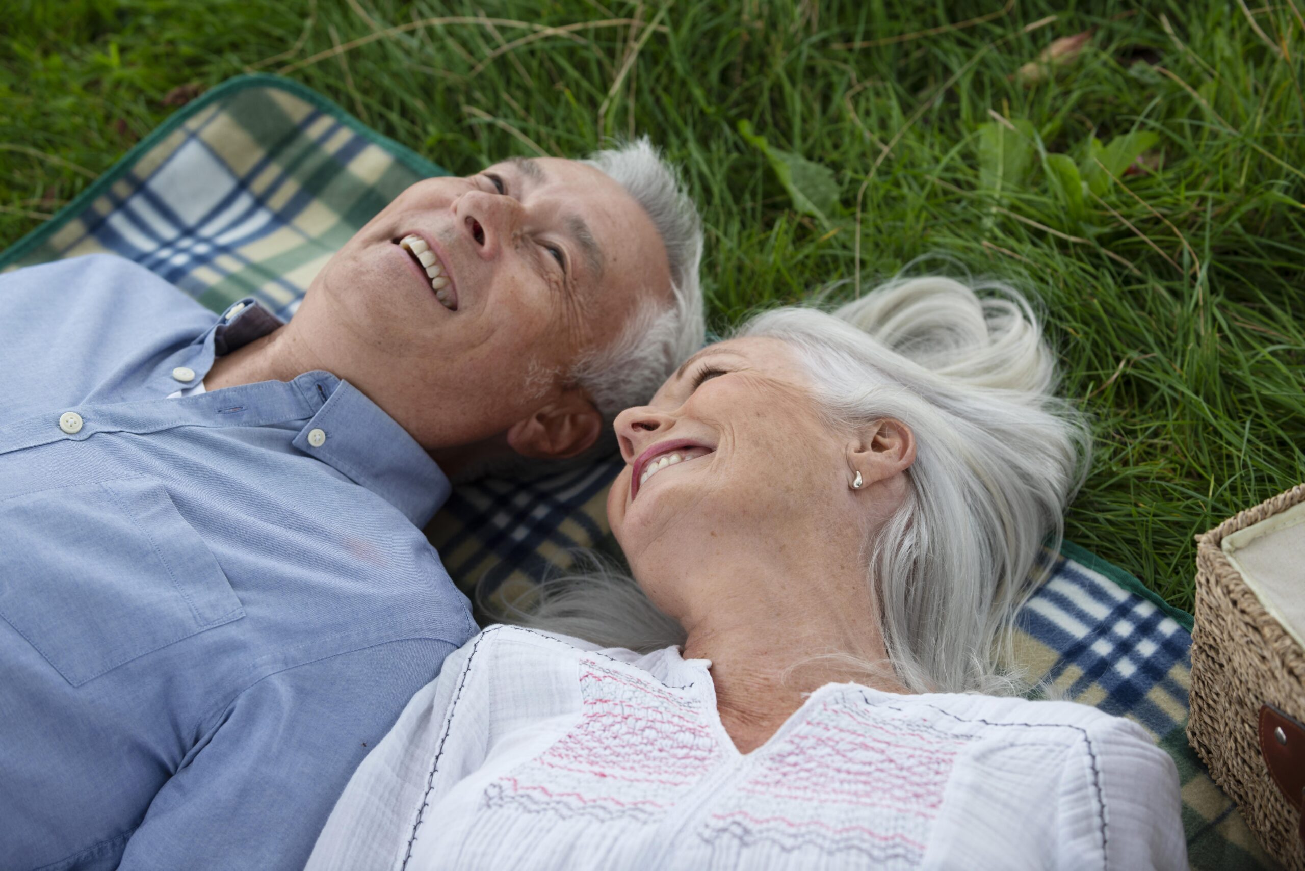 An older couple with grey hair lie on a chequered blanket in the grass, smiling and looking happy together. The woman rests her head close to the man as they chat about equity mortgage options, a picnic basket visible beside them.