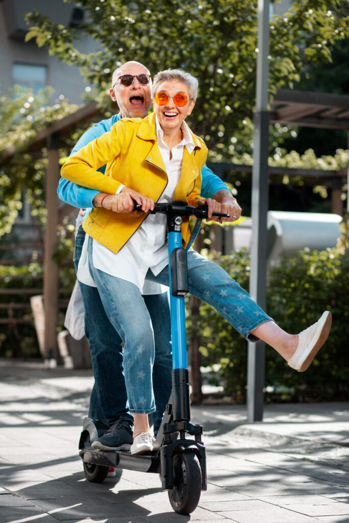 Two joyful pensioners ride together on an electric scooter outdoors; the woman in front smiles widely, wearing a yellow jacket and orange sunglasses, while the man behind her laughs, both enjoying themselves in a sunny park setting. Two joyful pensioners ride together on an electric scooter outdoors; the woman in front smiles widely, wearing a yellow jacket and orange sunglasses, while the man behind her laughs, both enjoying themselves in a sunny park setting.