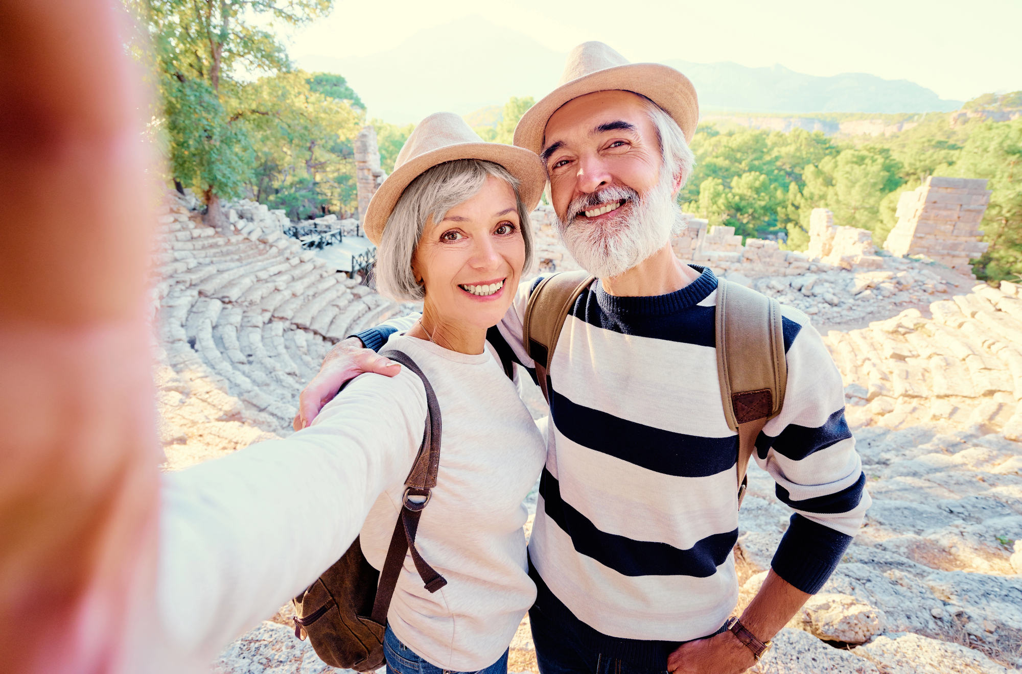 Smiling older couple wearing hats and rucksacks take a selfie together at ancient stone ruins, surrounded by sunlight and greenery—enjoying retirement adventures made possible by their equity release. Smiling older couple wearing hats and rucksacks take a selfie together at ancient stone ruins, surrounded by sunlight and greenery—enjoying retirement adventures made possible by their equity release.