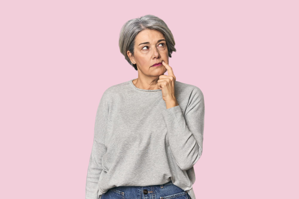 An older woman with short grey hair, the quintessential Silver Digital Nomad, stands against a pink background in a grey jumper and blue jeans, looking thoughtful with her finger resting on her chin. An older woman with short grey hair, the quintessential Silver Digital Nomad, stands against a pink background in a grey jumper and blue jeans, looking thoughtful with her finger resting on her chin.