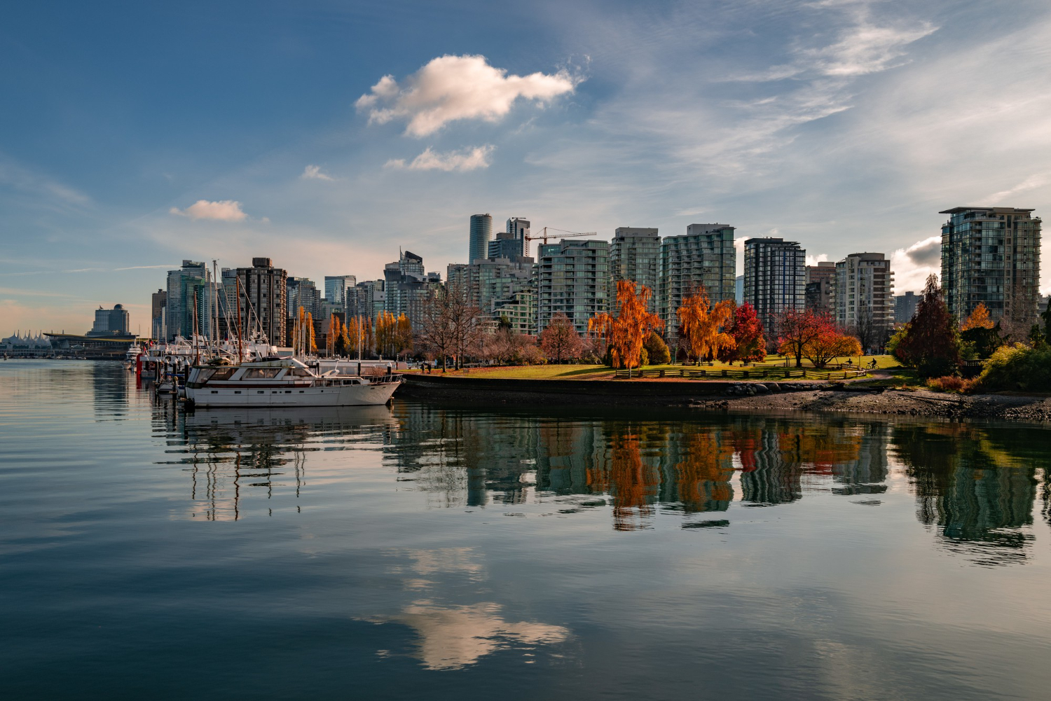 City centre Vancouver skyline with modern buildings, colourful autumn trees, and boats along the calm waterfront in our coverage area, all reflected in the water under a partly cloudy sky.