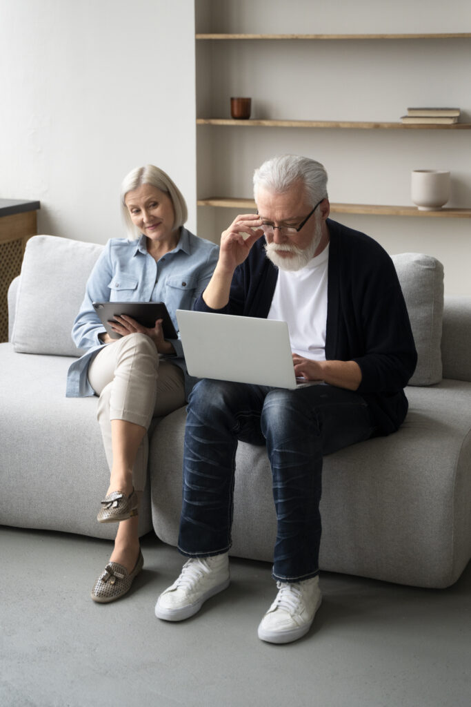 An older man and woman sit on a light sofa; the man uses a laptop while the woman, smiling, holds a tablet—perhaps checking news about inflation. Shelves with minimal décor are visible in the background. An older man and woman sit on a light sofa; the man uses a laptop while the woman, smiling, holds a tablet—perhaps checking news about inflation. Shelves with minimal décor are visible in the background.